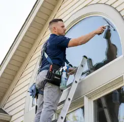 Professional window cleaner on a ladder using a squeegee to clean an exterior arched window, Oklahoma City, OK.